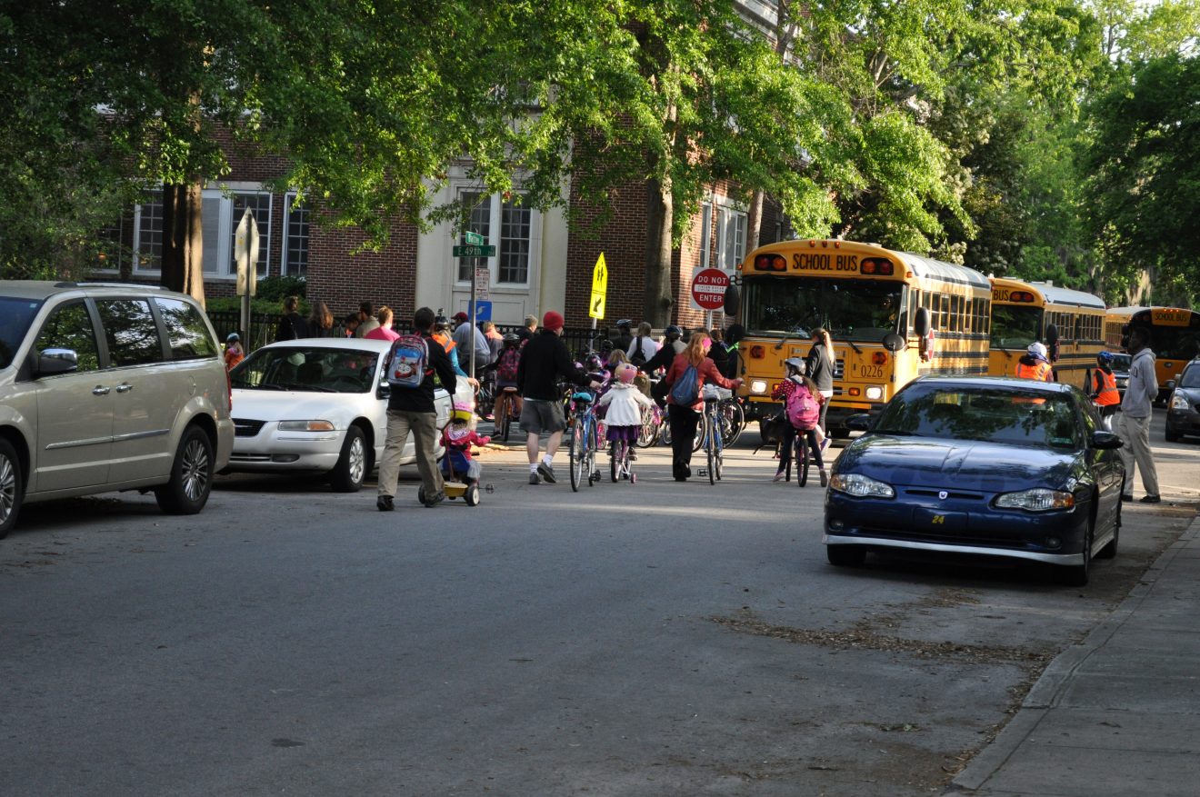 Children and parents walking and riding bikes to school. School bus dropping children off on corner of street.