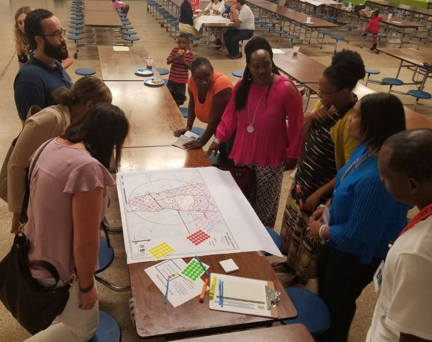 A group of parents, educators, and local professionals look over a map of the area around the school discussing concerns for safe walking and bicycling routes. Participants have access to dots and note pads as well as markers to mark the map with concerns and problem spots.