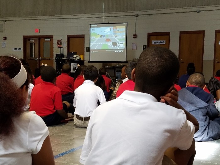Students sitting on the floor of a classroom watching a presentation on safe ways to commute to school.