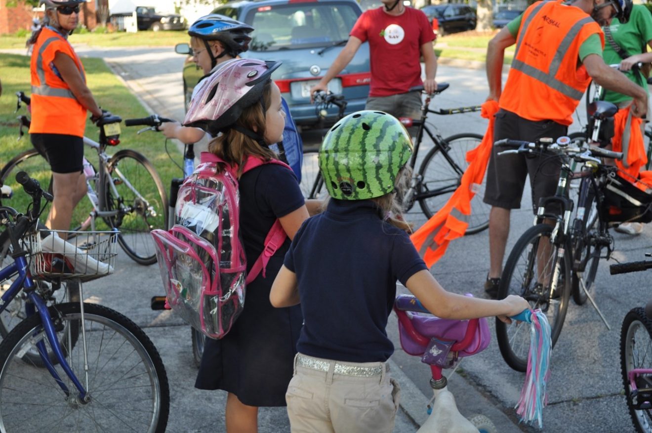 Families prepare to bike to school from a local park for a Bike to School Event in Savannah, Georgia. Two girls are holding their bikes in the foreground. One is wearing a watermelon helmet.