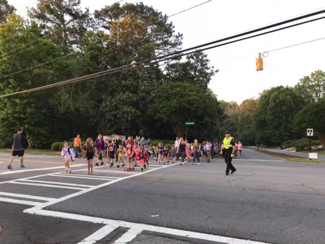 A large group of students and families cross an intersection with an officer in bright yellow vest directing traffic 